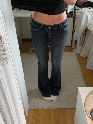 Mörkblå bootcut jeans med detaljer - Säljer ett par mörkblå bootcut jeans med snygga slitningar och kontrastsömmar. Bakfickorna har coola broderier och nitar som ger extra edge. Jeansen har låg midja och klassisk femficksmodell. Perfekta för dig som gillar en trendig och avslappnad stil.