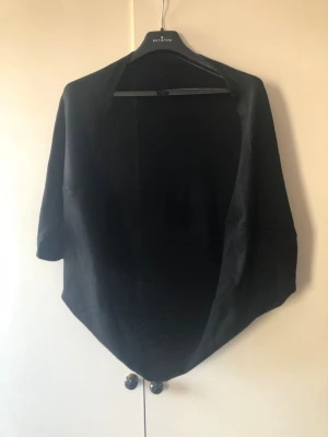 Svart Finstickad Cocoon-kofta / Cape – Onesize - Mångsidig och snygg svart finstickad kofta i draperad cocoon-/capemodell. Går ner över ryggen till rumpan, har vida ärmhål och är helt öppen framtill för ett fint fall. Perfekt över linnen och klänningar! Saknar material- och storlekslapp, men uppskattas som en mjuk viskos/akrylblandning. Flexibel i storleken och passar Onesize (ca S–L beroende på önskad passform). Fint begagnat skick!