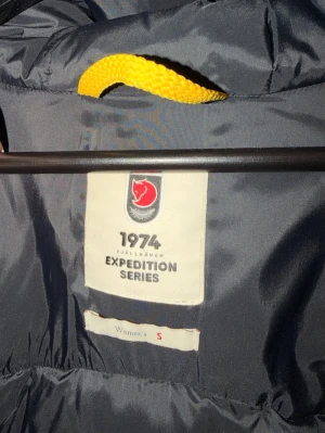 Fjällräven Expedition dunjacka S - Mörkblå dunjacka från Fjällräven Expedition Series 1974 med gul hängare i nacken. Jackan är lätt och har klassisk design med hög krage. Perfekt för kalla vinterdagar tack vare dunfyllningen som håller värmen.
