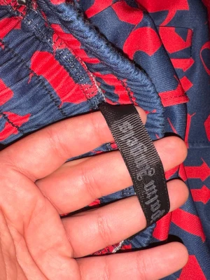 Blå-röda mjukisbyxor med tryck - Säljer ett par blå mjukisbyxor med rött grafiskt tryck och röda sidoremsor. Byxorna har resår i midjan och raka ben. Materialet ser ut att vara polyester eller liknande för en sportig känsla. Perfekta för dig som gillar streetwear och vill sticka ut.