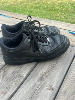 Svarta air force 1 - Säljer ett par helsvarta af1 med lågt skaft och klassisk siluett. Skorna har svarta skosnören, perforerad tå och detaljer i svart läder. Passar perfekt till streetwear och vardagsstil.