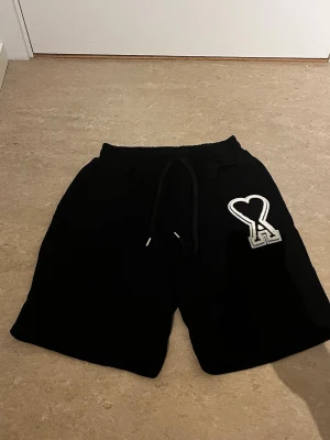 Svarta shorts med hjärtdetalj - Snygga svarta shorts med resår och dragsko i midjan. På vänster ben finns en stor vit patch med ett hjärta och bokstaven A. Perfekta för en avslappnad och trendig look. Materialet ser ut att vara mjukt och bekvämt, troligen bomull eller bomullsblandning.