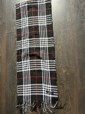 Rutig halsduk från Burberry i kashmir - Säljer en klassisk halsduk från Burberry i 100% kashmir. Halsduken har svart bas med vitt och rött rutmönster samt fransar i ändarna. Tillverkad i England och supermjuk, perfekt för att lyfta din outfit.