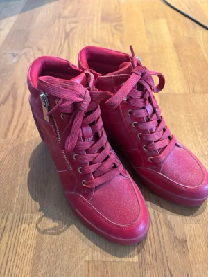Röda high top sneakers med kilklack - Snygga röda high top sneakers med kilklack, dragkedja på sidan och snörning framtill. Skorna har detaljer i både textil och glansigt material samt guldfärgade metalldetaljer vid dragkedjan och hälen. Perfekta för dig som vill sticka ut med din stil.