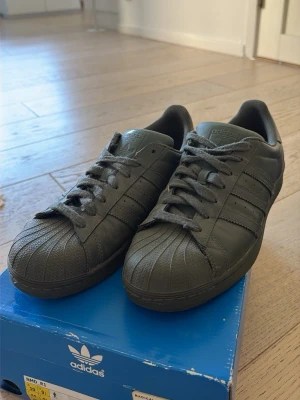 Mörkgröna Adidas Superstar sneakers - Säljer ett par mörkgröna Adidas Superstar sneakers i läder med klassisk snäcktå och de ikoniska tre ränderna på sidan. Pharrell Williams collab. 