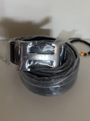 Svart H bälte - Snyggt svart bälte i skinn med ett stort, blankt silverspänne format som ett H. Perfekt accessoar för att lyfta din outfit och ge en clean look. Bältet har klassisk design och passar till både jeans och kostymbyxor.