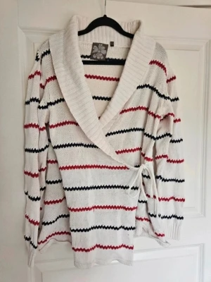 Omlottkofta Pippi XL - Omlottkofta Pippi XL Stickad omlottkofta i vitt med blå och röda horisontella ränder. Koftan har en bred, ribbad sjalkrage och knyts i sidan med ett band. Långärmad och i mjukt material, perfekt för lager på lager och en avslappnad look.  Välanvänd!  Kan även hämtas I Partille  E1