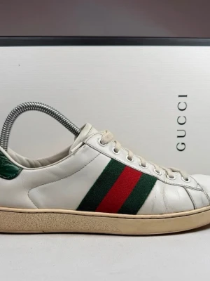 Gucci vita sneakers med röd/grön rand - Klassiska vita sneakers från Gucci med den ikoniska gröna och röda randen på sidan. Skorna har lågt skaft, vita snören och en beige sula. Materialet är slätt läder med detaljer i grönt på hälen. Perfekta för dig som gillar stilrena och lyxiga sneakers. Priset är ej hugget i sten så kom med bud🤝