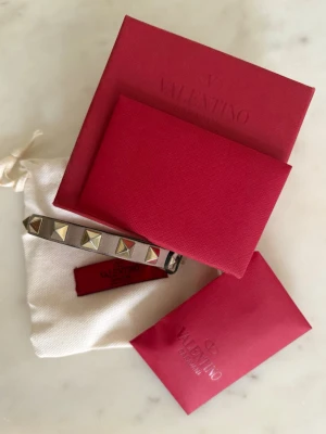 Valentino läderarmband med nitar - Snyggt armband från Valentino i rosa/beige läder med ikoniska pyramidformade nitar. Levereras med originalförpackning, dustbag och två extra oanvända nitar. Varan har en liten defekt i slutet av lädret, men ingenting som märks under användning. 