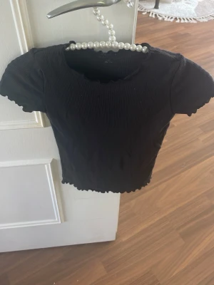 Svart ribbad croppad topp - Säljer en svart ribbad croppad topp med korta ärmar och vågig kant vid ärmslut och nederkant. Perfekt till jeans eller kjol för en enkel men snygg look.