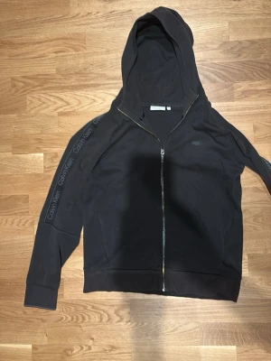 Svart hoodie med dragkedja Calvin Klein - Svart hoodie från Calvin Klein med dragkedja framtill och huva. Långa ärmar med Calvin Klein-logga längs axlarna och ärmarna. Tillverkad i mjukt material, perfekt för en avslappnad och stilren look.