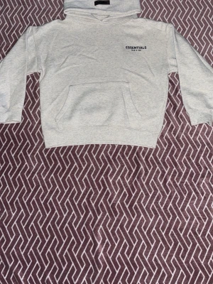 Grå Essentials Fear of God hoodie - Snygg ljusgrå hoodie från Essentials Fear of God med svart logga på bröstet och stor text på ryggen. Klassisk känguruficka framtill och huva. Perfekt för en clean och avslappnad streetwear-look.   Pris kan diskuteras 