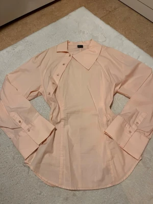 Ljusrosa skjorta från Gina Tricot - Säljer en ljusrosa skjorta från Gina Tricot med klassisk krage och långa ärmar. Skjortan har knappar framtill och breda manschetter med knappdetaljer. Perfekt att styla till jeans eller kjol för en clean look.