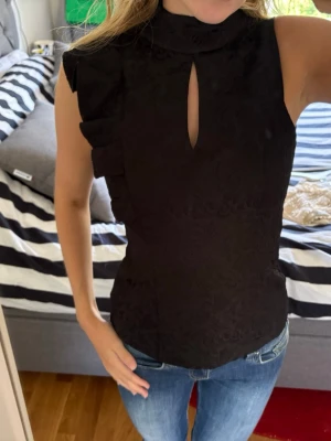 Svart ärmlös topp med volangdetaljer - Säljer en svart, ärmlös topp med hög krage och snygg öppning framtill. Toppen har volangdetaljer längs axlarna och ett diskret mönster i tyget som ger en lyxig känsla. Passformen är figurnära och toppen är perfekt till jeans eller kjol.