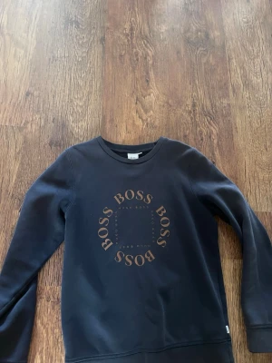 Svart sweatshirt från BOSS - Snygg svart sweatshirt från BOSS med rund hals och tryck i form av cirkulär logga på bröstet. Tröjan har långa ärmar och är tillverkad i mjukt material som passar perfekt till en avslappnad stil.