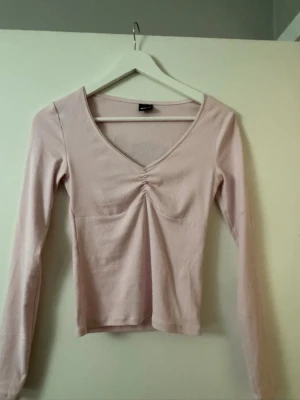 Ljusrosa långärmad topp med rynk - Säljer en ljusrosa långärmad topp med v-ringning och snygg rynkdetalj framtill. Toppen har en slim passform och är gjord i mjukt bomullsmaterial. Perfekt att styla med jeans eller kjol för en enkel men söt look.