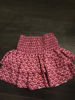 Rosa mönstrad volangkjol - Kort volangkjol från Meet x Tiger i rosa och rött med smock i midjan. Kjolen har ett grafiskt mönster och dubbla volanger som ger en lekfull look. Perfekt för dig som vill ha en färgstark och trendig kjol. Med inbyggda shorts.