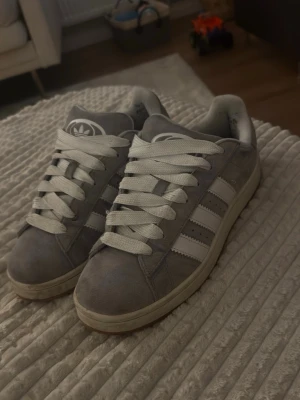 Adidas - Snygga grå sneakers från Adidas med klassiska vita ränder på sidorna och vit sula. Skorna har breda vita skosnören och är tillverkade i mockaliknande material. Perfekta för en stilren och sportig look.
