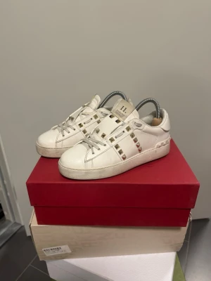 Vita sneakers med nitar - Snygga vita sneakers med coola guldfärgade nitar längs sidorna och över vristen. Skorna har klassisk låg modell med vita snören och platt sula. Tillverkade i läder för en stilren och lyxig känsla. Perfekta för dig som vill sticka ut med detaljer. Ingen box ingår