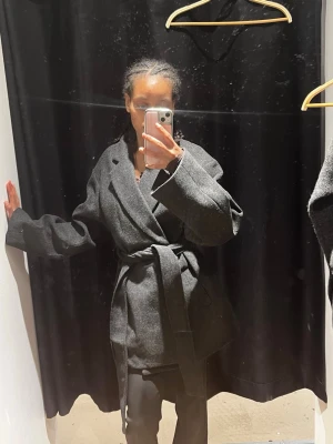 Mörkgrå omlottkappa med skärp - Snygg mörkgrå kappa i omlottmodell med brett skärp i midjan och stora slag. Kappan har en rak passform och långa ärmar, perfekt för dig som gillar en stilren och trendig look. Materialet ser ut att vara ullblandning och färgen är klassiskt mörkgrå.