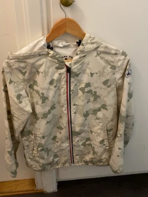 vindjacka med huva - Snygg vindjacka i ljus camouflage-mönster med dragkedja framtill och huva. Jackan har två fickor och en patch på ärmen. Perfekt för dig som vill ha en streetig och sportig look. Materialet är tunt och smidigt, passar bra till lager-på-lager.