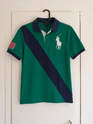 Ralph Lauren piké - Snygg och klassisk Ralph Lauren-piké i grön färg med marinblå detaljer och stort broderat Polo-emblem på bröstet. Tröjan har även en amerikansk flagga på ärmen, vilket ger den en unik och sportig look. ✓ Original Ralph Lauren ✓ Grön med marinblå detaljer ✓ Broderat emblem framtill ✓ Bra skick ✓ Perfekt till både vardag och sommar