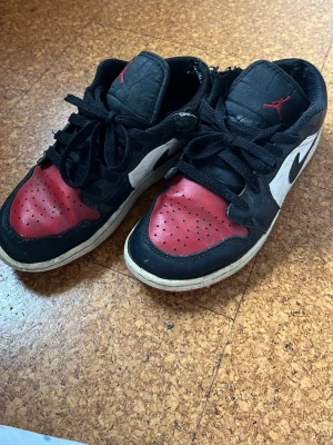 Svarta och röda Nike Air Jordan 1 Low - Säljer ett par Nike Air Jordan 1 Low sneakers i svart, rött och vitt. Skorna har klassisk låg silhuett, svart swoosh på sidan och röd tåbox. Tillverkade i läder med svarta skosnören och vit mellansula. Perfekta för dig som gillar streetwear och ikoniska sneakers.Skorna visar tydliga tecken på användning men de är äkta 