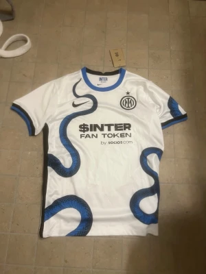 Inter Milan vit matchtröja Nike - Säljer en vit Inter Milan matchtröja från Nike med blå och svarta ormmönster, svart logga och sponsortryck på bröstet. Tröjan har rund hals med blå kant och korta ärmar med blå detaljer. Perfekt för dig som älskar fotboll och vill sticka ut på planen.