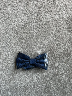 Mörkblå mönstrad fluga - Snygg mörkblå fluga med diskret blommönster i satinliknande material. Perfekt accessoar för att lyfta din outfit med en elegant touch. Flugan är förknuten och har ett klassiskt utseende.