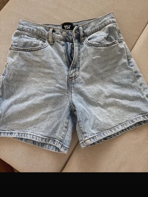 Ljusblå denimshorts från 157 - Säljer ett par ljusblå denimshorts från 157 med klassisk femficksdesign och normal passform. Shortsen har bälteshällor, knapp och dragkedja framtill samt två bakfickor. Perfekta för varma dagar och enkel att matcha med allt.