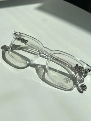 Chrome hearts - Snygga glasögon med transparent båge och rektangulär form. Bågarna har metalliska detaljer vid gångjärnen och ett diskret blommönster på ena linsen. Perfekt accessoar för dig som vill sticka ut med en modern och stilren look.