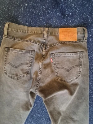 Grå Levi's 501 raka jeans - Säljer ett par klassiska grå Levi's 501 jeans med raka ben och femficksmodell. Jeansen har den ikoniska läderpatchen bak i midjan och röd Levi's-tagg på ena bakfickan. Tillverkade i slitstark denim med vintagekänsla och snygg tvätt. Det är lite slitna längst ner på byxbenna men funkar utmärkt ändå. 30 30
