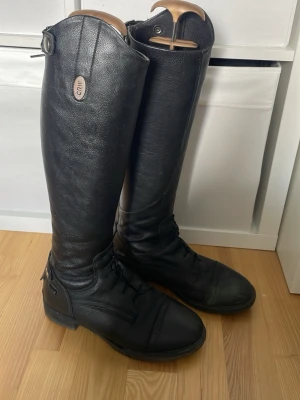 Svarta ridstövlar läder CRW - Kommer med guldiga stövelsträclare från hööks. Snygga svarta ridstövlar från CRW i äkta läder. Höga med klassisk form, snörning framtill och dragkedja bak. Perfekta för dig som vill ha en stilren look i stallet eller till ridningen. Metallmärke med CRW-logga upptill och justerbart spänne vid skaftet.