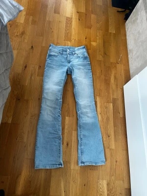 Ljusblå bootcut jeans - Säljer ett par ljusblå bootcut jeans med klassisk femficksdesign och utsvängda ben. Jeansen har slitningar framtill för en trendig look och är tillverkade i denim med stretch. Perfekta för dig som gillar retrovibbar och vill ha ett par bekväma jeans med snygg passform.