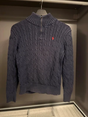 Mörkblå kabelstickad tröja Ralph Lauren - Snygg mörkblå kabelstickad tröja från Ralph Lauren med halv dragkedja och hög krage. Tröjan har det klassiska röda Polo-logotypen broderad på bröstet och ribbade muddar vid ärmslut och nederkant. Perfekt för en stilren och avslappnad look. Ett hål i armen som enkelt går att fixa snyggare om man kan sy lite.
