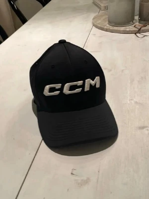 Svart keps från CCM med brodyr - Snygg svart keps från CCM med stor vit broderad logga framtill. Kepsen har böjd skärm och ventilerande mesh på baksidan för extra komfort. Perfekt för dig som gillar sportig stil och vill ha en klassisk look.