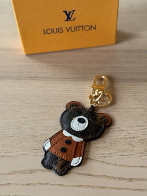 Vuitton björn nyckelring - Säljer en söt nyckelring från  Vuitton i form av en björn. Nyckelringen är gjord i brunt, svart och vitt läder med guldfärgad metallhake. Perfekt att hänga på väskan eller nycklarna för en lyxig touch.