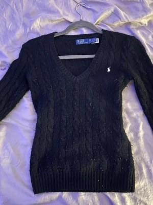 Svart kabelstickad tröja Polo Ralph Lauren - Svart kabelstickad tröja från Polo Ralph Lauren med v-ringning och vit broderad logga på bröstet. Tröjan har lång ärm och ribbade muddar vid ärmslut och nederkant. Perfekt för lager-på-lager och snygg till jeans eller kjol. 20% kashmir 80% ull, köpt i New york 2024 men knappt använd💞