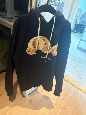 Svart hoodie med nalle från Palm Angels - Svart hoodie med stor broderad nalle på bröstet och vita dragsnören i huvan. Klassisk känguruficka framtill och avslappnad passform. Perfekt för dig som gillar streetwear och vill sticka ut med en unik detalj.