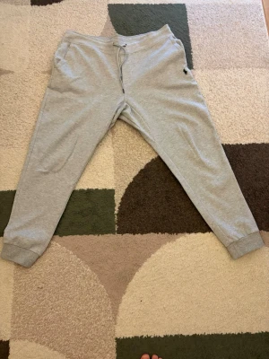 Grå mjukisbyxor med dragsko - Säljer ett par grå mjukisbyxor fråm ralph lauren, med dragsko i midjan och muddar vid bensluten. Byxorna har fickor på sidorna och en bakficka. Perfekta för chill dagar hemma eller när du vill vara bekväm. Materialet känns mjukt och skönt mot huden.