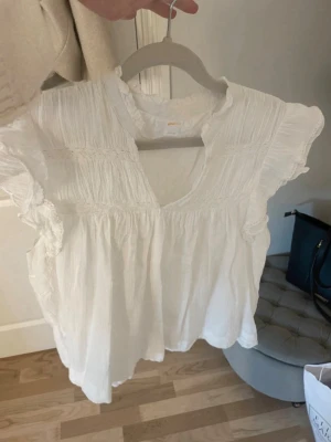 Vit blus från Gina Tricot - Söt vit blus från Gina Tricot med volangdetaljer och rynkade partier. Blusen har en lös passform, v-ringning och korta ärmar med volang. Perfekt för en fräsch look. Tillverkad i ett lätt och luftigt tyg.
