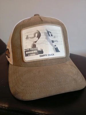 Beige och vit keps med Daffy Duck - Säljer en beige och vit keps med mesh på sidorna och ett stort tygmärke framtill med Daffy Duck och ACME Police-tema. Kepsen har Looney Tunes-detalj på sidan och böjd skärm. Perfekt för dig som gillar tecknade klassiker och streetwear. Nypris 500 kr. Nästan oanvänd men lite smutsig på insidan (se bild 3). Pris kan diskuteras🙌. Mvh Rasmus.