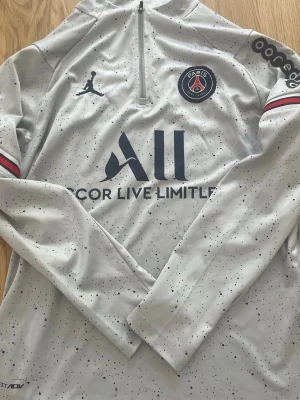 PSG träningströja ljusgrå långärmad - Snygg långärmad träningströja från PSG i ljusgrått med svarta prickar, halv dragkedja och klubbmärke på bröstet. Röd och svart detalj på ärmarna och tryck på både fram- och baksida. Tillverkad i ett tekniskt funktionsmaterial som passar perfekt till träning.