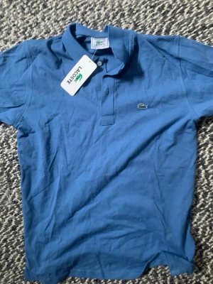 Blå pikétröja från Lacoste - Säljer en klassisk blå pikétröja från Lacoste med den ikoniska krokodilloggan på bröstet. Tröjan har korta ärmar, krage och knappar framtill. Perfekt för en clean och sportig stil.