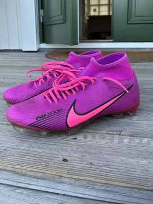  Nike fotbollsskor - Säljer ett par lila Nike fotbollsskor med högt skaft och rosa detaljer. Skorna har snörning, tydlig Nike-logga på sidan och dobbar för bra grepp på planen. Tillverkade i syntetmaterial och designade för fart och kontroll på konstgräs eller naturgräs.