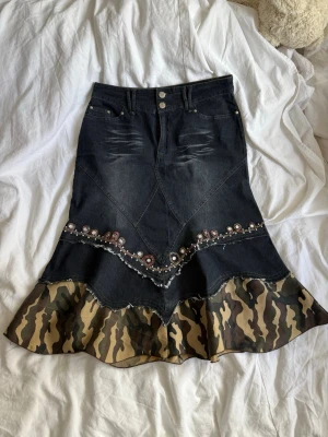 Jeanskjol med camouflagevolang - Unik jeanskjol med dekorativa pärlor och broderier samt volang i camouflage-tyg längst ner. Kjolen har hög midja, knappstängning och coola sömdetaljer. Perfekt för dig som vill sticka ut med din stil.