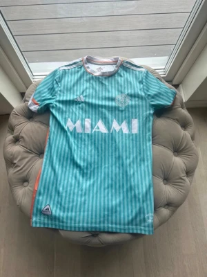 Turkos randig Miami matchtröja Adidas - Säljer en turkos och vitrandig matchtröja från Adidas med MIAMI-tryck på bröstet. Tröjan har vita och orange detaljer, rund hals och är tillverkad i lätt och ventilerande AEROREADY-material. Perfekt för fotboll eller träning.