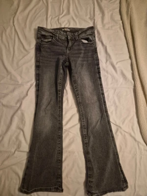 Grå bootcut jeans med fickdetaljer - Säljer ett par grå bootcut jeans med snygga slitningar och klassiska fem fickor. Jeansen har knapp- och dragkedjestängning samt dekorativa ficklock med knappar bak. Säljer dessa för att dem inte längre passar. Storlek 158 från gina tricot