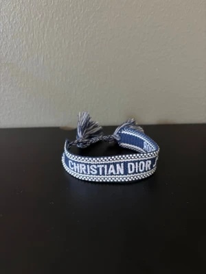 Blått Christian Dior armband - Snyggt blått armband från Christian Dior med vit brodyr och text. Armbandet har dekorativa tofsar i ändarna och är gjort i textilmaterial. Perfekt accessoar för att lyfta din outfit med en exklusiv touch..                                Använd någon gång endast! Har ej kvitto! 