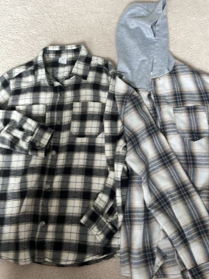 Skjortor 👔  - Säljer två overshirts i mjuk flanell. Ena är svartvit-rutig med klassisk krage och bröstficka, den andra är beige-blå-rutig med grå huva och bröstficka. Perfekta att slänga över en t-shirt för en avslappnad look.